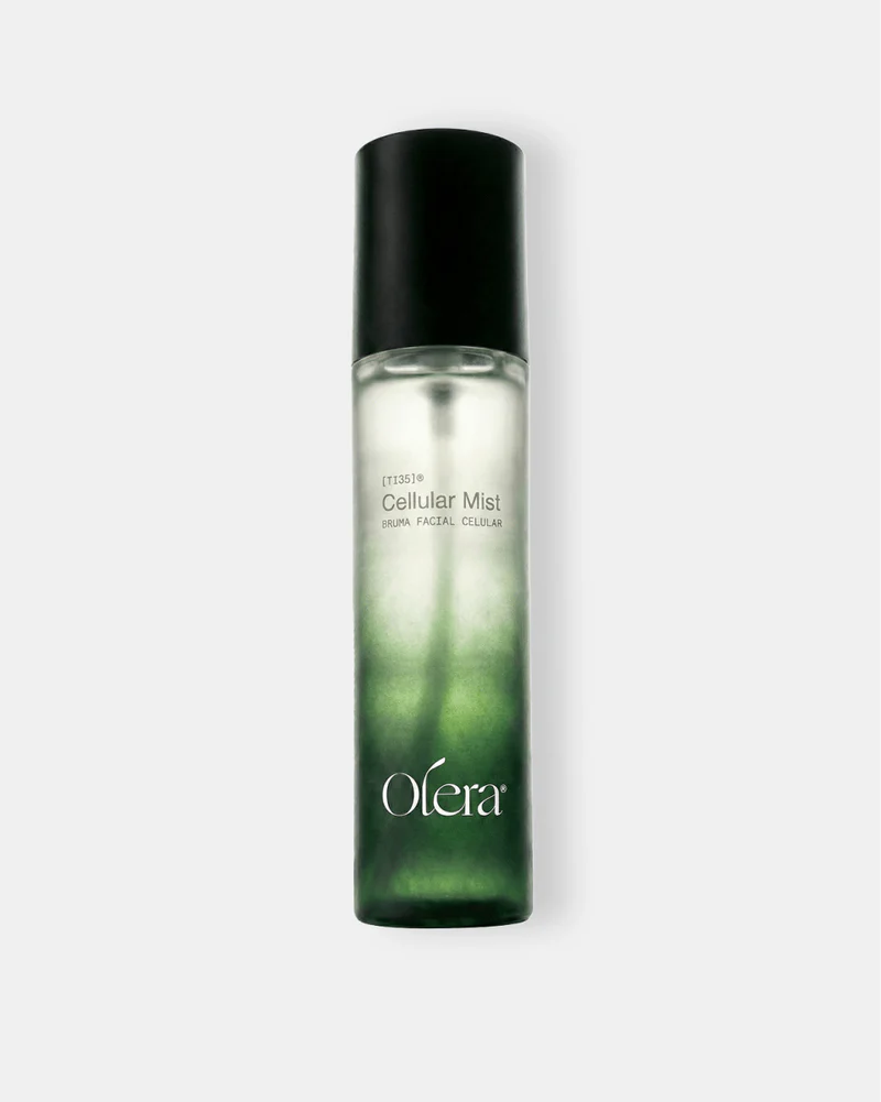 Frasco de spray de vidro transparente com tampa preta, contendo um l&iacute;quido que transita do verde escuro na base para o transl&uacute;cido no topo. O r&oacute;tulo branco exibe Cellular Mist BRUMA FACIAL CELULAR e o logo Olera em branco