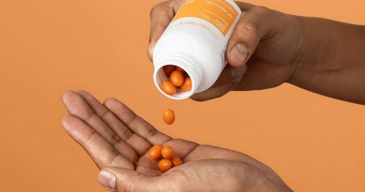 M&atilde;o de pele escura despejando c&aacute;psulas alaranjadas de um frasco branco em outra m&atilde;o aberta, contra fundo laranja