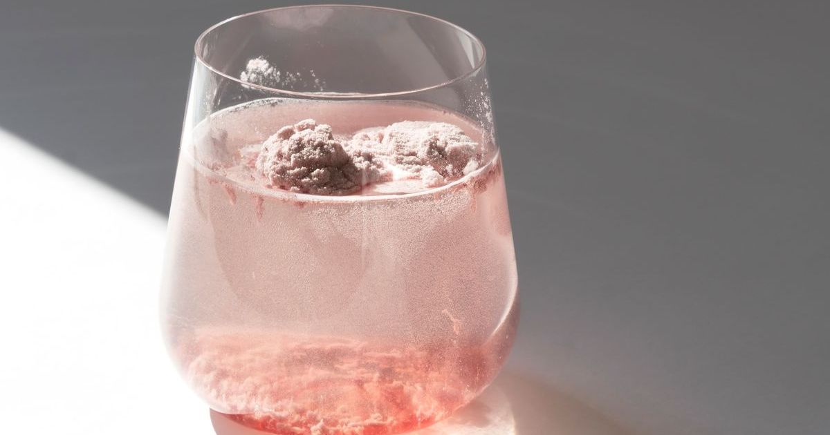 Um copo transparente com uma bebida rosa efervescente, com duas bolas de p&oacute; rosa flutuando na superf&iacute;cie e sedimentos avermelhados no fundo, sobre um fundo cinza com uma sombra diagonal