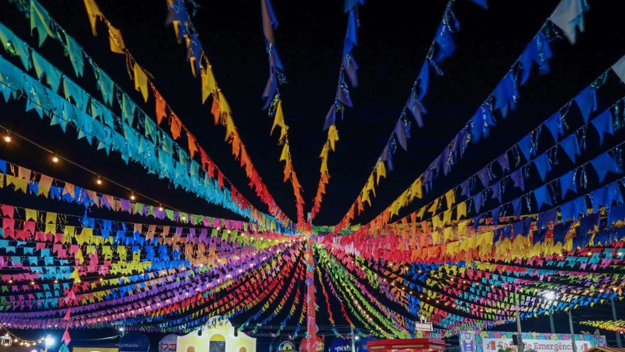 Céu noturno escuro com centenas de bandeirinhas coloridas de festa junina, em tons de azul, amarelo, vermelho, rosa e verde, penduradas em cordões que convergem para o centro. Pequenas lâmpadas incandescentes brilham entre as bandeirinhas, iluminando o ambiente festivo. Ao fundo, estruturas de barracas e decorações de festa junina são visíveis, com uma placa branca e vermelha indicando "Sala de Emergência" à direita