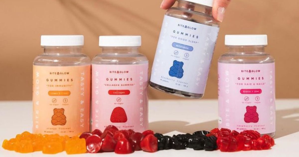 Quatro potes de vitaminas em goma Bite & Glow, com r&oacute;tulos laranja, rosa claro, azul claro e rosa escuro, e gomas coloridas espalhadas na frente. Uma m&atilde;o segura o pote azul claro.