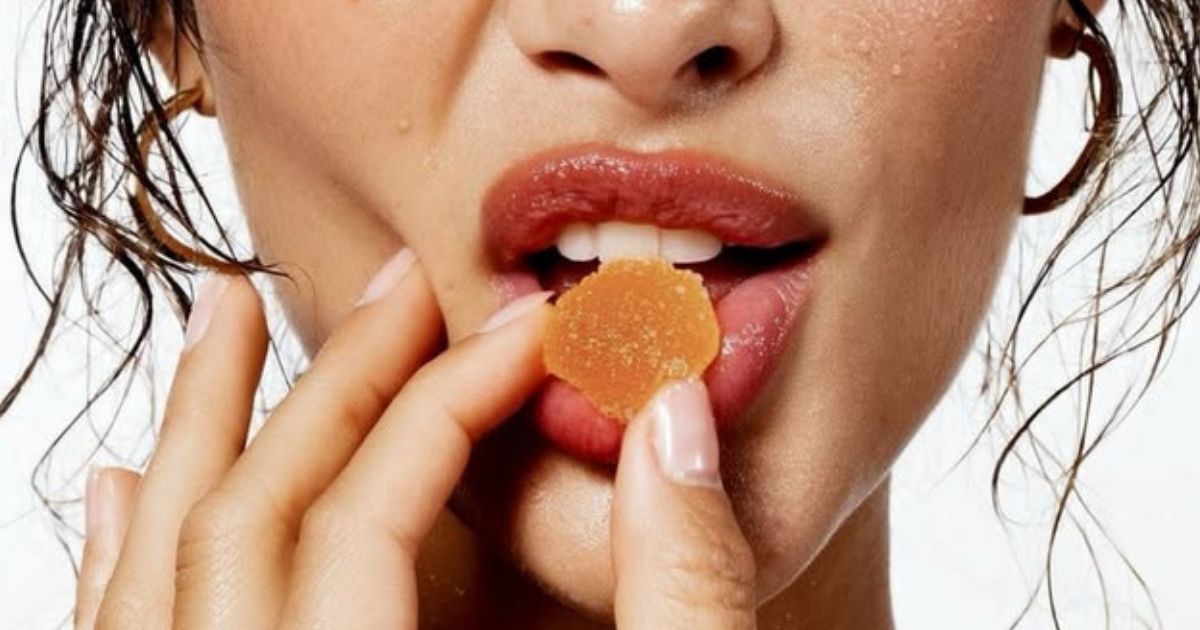 Close-up de uma mulher com pele suada e cabelos molhados, segurando uma goma de cor laranja com os dedos e levando-a à boca com os lábios entreabertos
