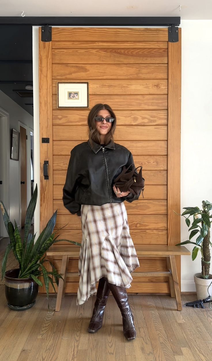 Mulher sorridente de &oacute;culos escuros, jaqueta preta, saia xadrez bege e botas marrons, segurando uma bolsa marrom, em frente a uma porta de madeira com plantas ao redor