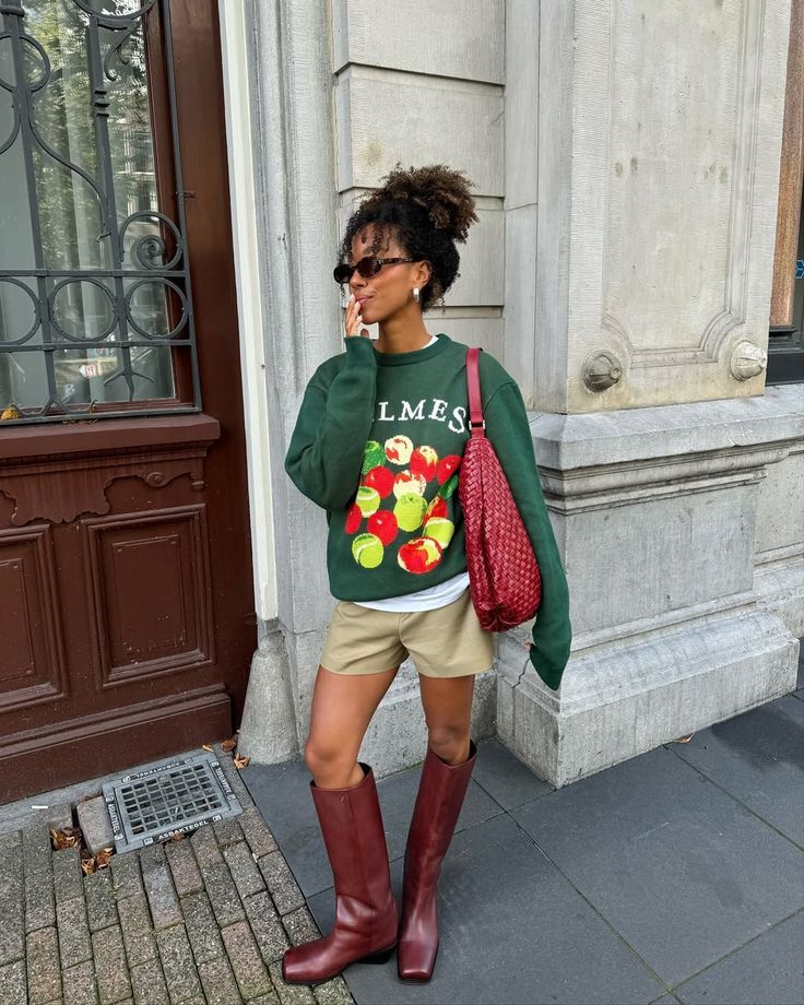 Mulher de pele morena, cabelo crespo preso no alto, &oacute;culos escuros e brincos de argola, vestindo um moletom verde com estampa de bolas coloridas, shorts c&aacute;qui e botas de cano alto cor vinho, segurando uma bolsa vermelha tran&ccedil;ada no ombro, em p&eacute; na cal&ccedil;ada de um pr&eacute;dio com fachada de pedra e porta de madeira