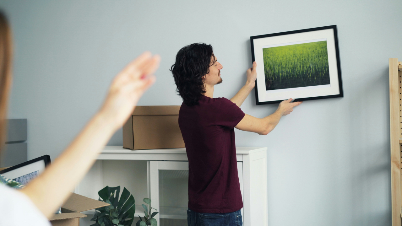 Homem de cabelo escuro e camiseta vinho pendura um quadro com imagem de campo verde em parede azul clara, enquanto uma mulher com braço estendido o orienta. Caixas de mudança e plantas decoram o ambiente