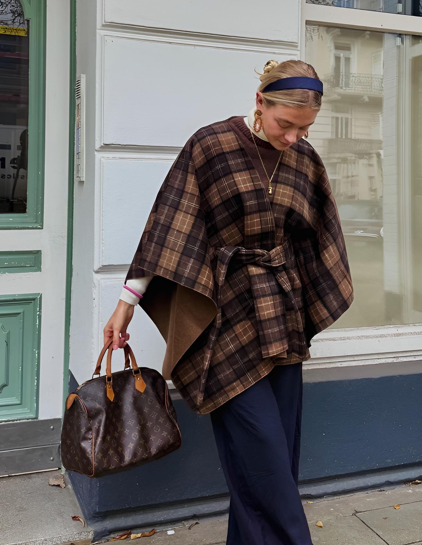 Mulher loira com coque e tiara azul, usando capa xadrez marrom e bege, blusa branca, cal&ccedil;a azul marinho, brincos dourados e colar, segurando bolsa Louis Vuitton marrom, em frente a uma fachada branca com porta verde