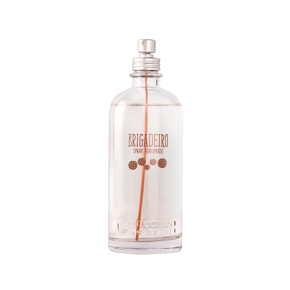 Frasco transparente de spray perfumado Brigadeiro da LOccitane Au Br&eacute;sil, com l&iacute;quido claro e tubo interno laranja. O r&oacute;tulo tem letras em tom cobre e desenhos de granulados de brigadeiro