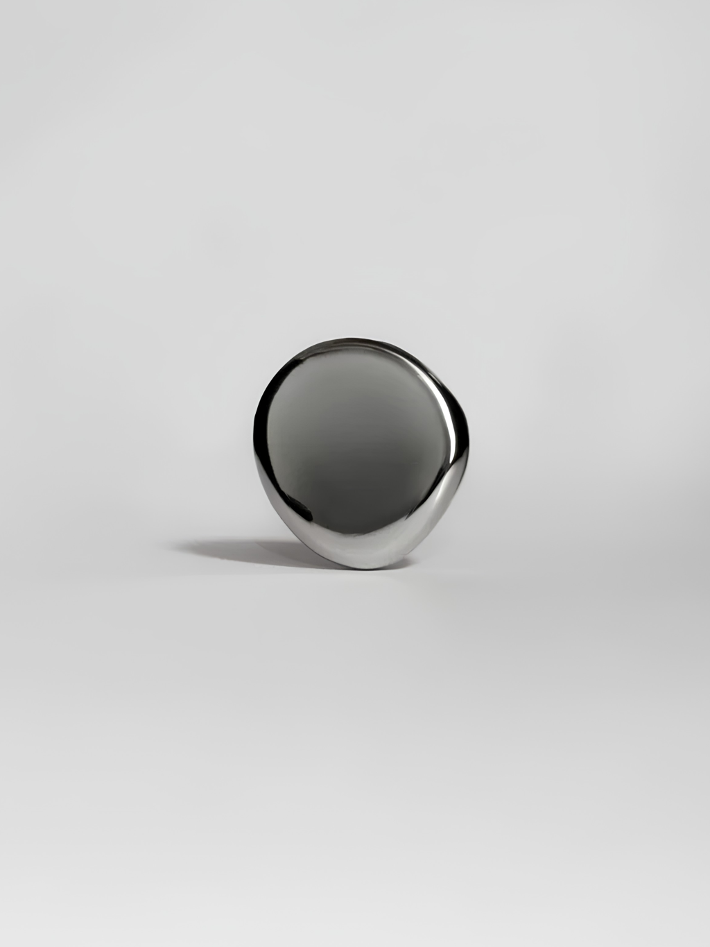 Objeto met&aacute;lico prateado, liso e brilhante, com formato oval irregular, apoiado em uma superf&iacute;cie branca. Uma sombra escura e alongada se projeta &agrave; esquerda, criando contraste e profundidade. A ilumina&ccedil;&atilde;o destaca o reflexo na superf&iacute;cie do objeto.