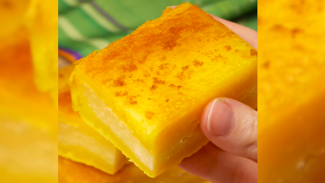 Mão segurando um pedaço de bolo de fubá cremoso, com a superfície dourada e algumas manchas mais escuras, revelando camadas claras no interior. Ao fundo, um tecido verde desfocado
