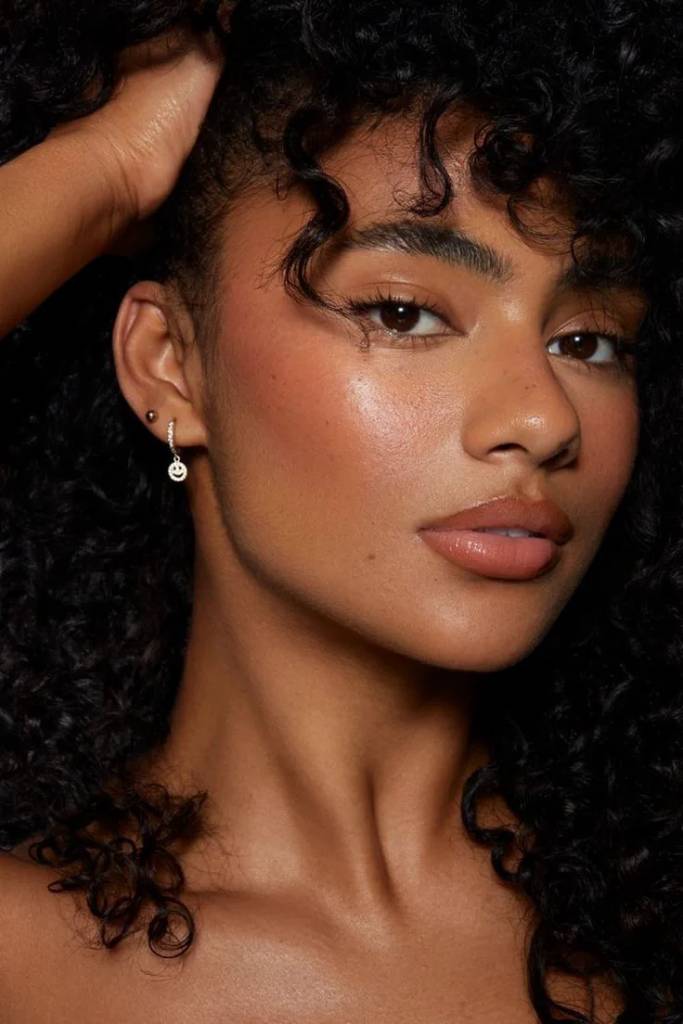 Mulher jovem com pele morena e cabelos cacheados escuros, olhando diretamente para frente. Ela tem um brinco de argola com um rosto sorridente e um brinco pequeno na orelha esquerda. Sua m&atilde;o direita est&aacute; levantada, com os dedos entre os cachos do cabelo. Ela usa maquiagem leve, com blush nas ma&ccedil;&atilde;s do rosto e batom nude nos l&aacute;bios. O fundo &eacute; escuro, destacando seu rosto e cabelo.