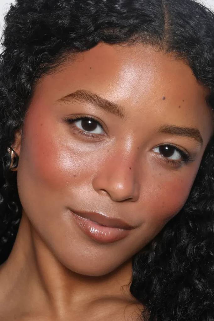 Rosto de uma mulher jovem com pele morena, cabelo cacheado escuro, maquiagem natural com blush rosado e l&aacute;bios brilhantes, olhando para frente com um leve sorriso