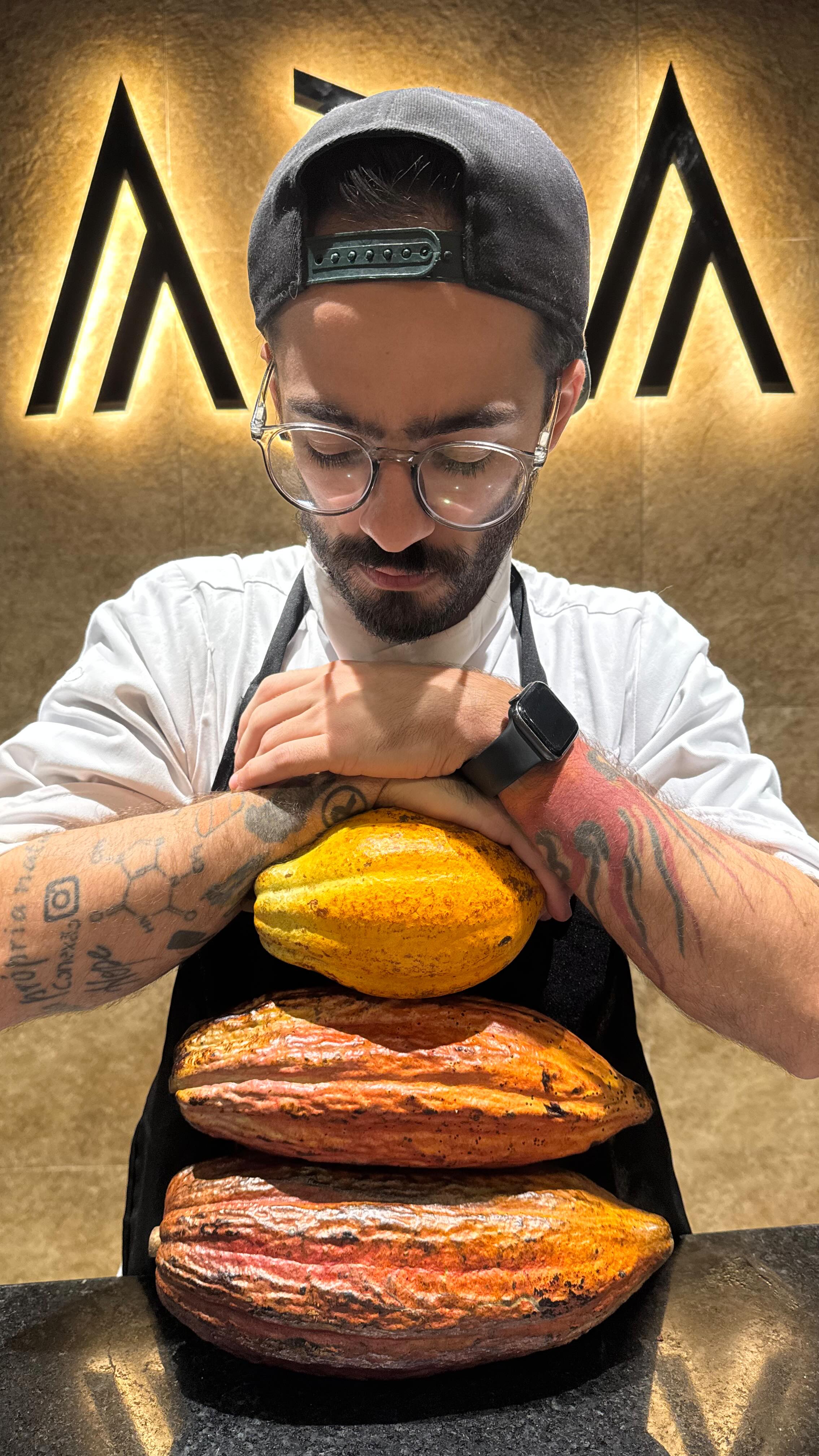 Homem barbudo de &oacute;culos, bon&eacute; e avental preto, com tatuagens nos bra&ccedil;os, apoia os bra&ccedil;os sobre tr&ecirc;s frutos de cacau empilhados, olhando para baixo. Ao fundo, um logo iluminado com duas letras A estilizadas