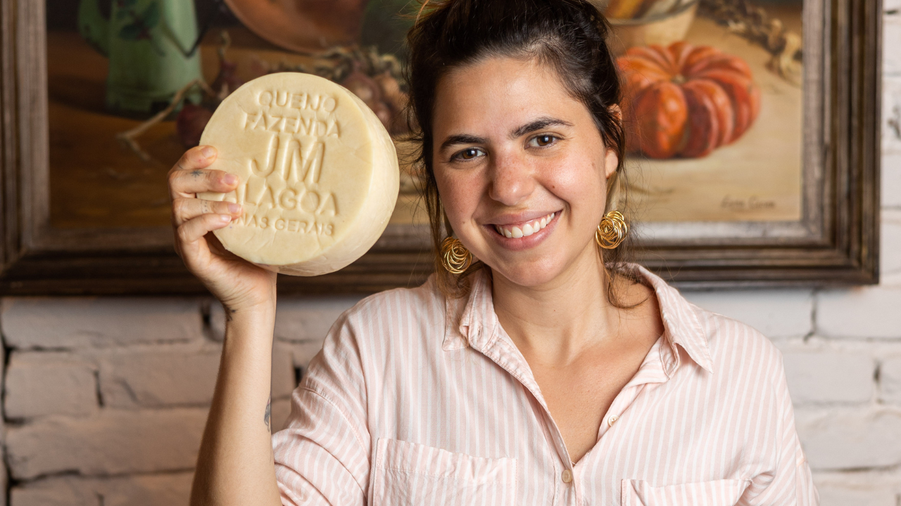 Mulher sorridente, de camisa listrada clara e brincos dourados, segura um queijo redondo com a marca QUEIJO FAZENDA JM LAGOAS MINAS GERAIS. Ao fundo, uma pintura de abóbora e uma parede de tijolos brancos