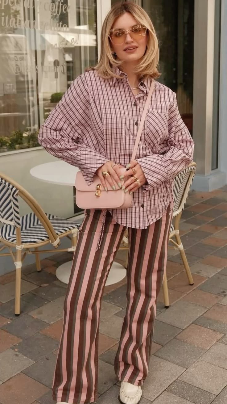 Mulher loira de &oacute;culos escuros e blusa xadrez rosa e branca, cal&ccedil;a listrada rosa e marrom, bolsa rosa claro, posando em frente a um caf&eacute; com cadeiras de vime