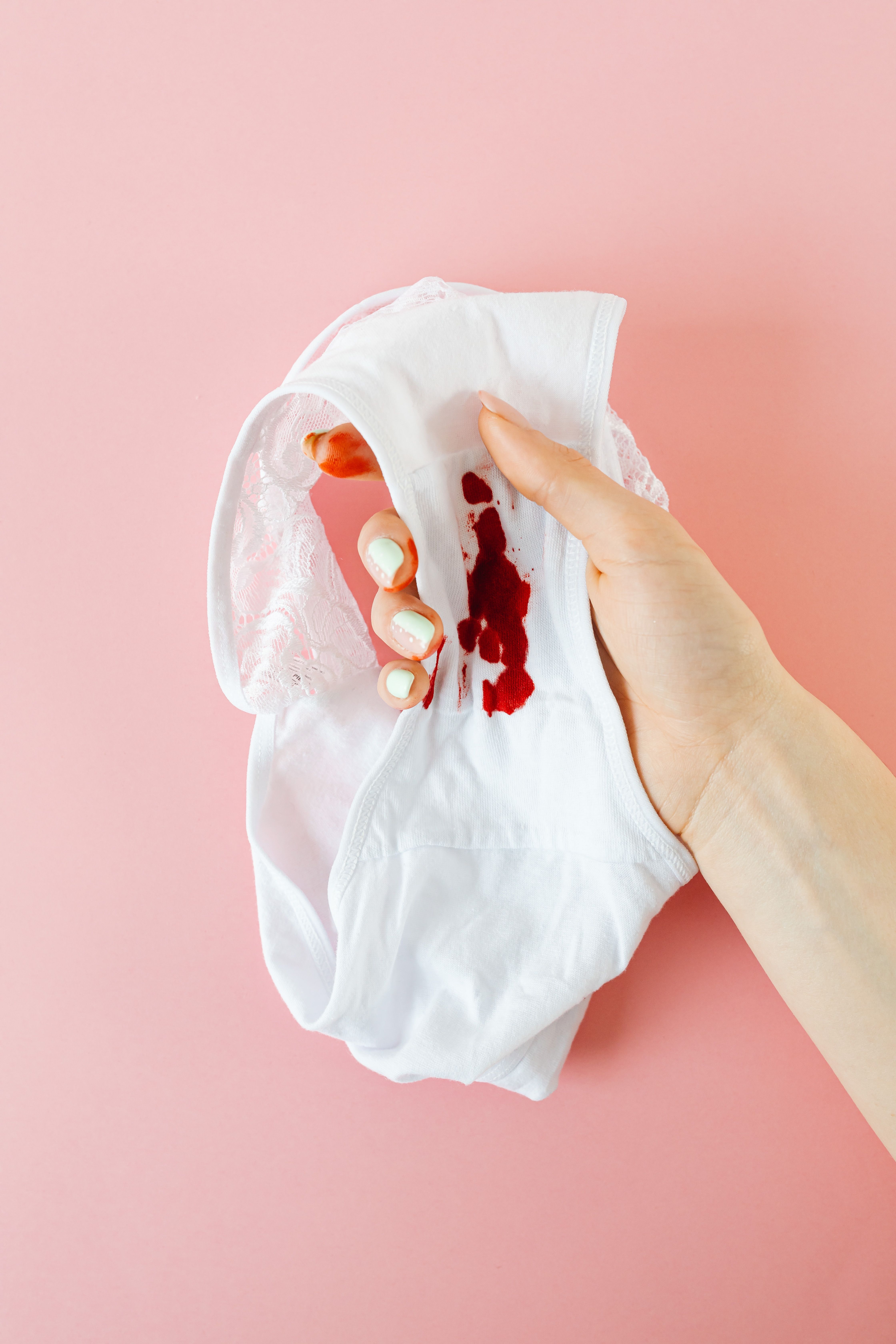 M&atilde;o feminina segura calcinha branca com mancha de sangue menstrual, unhas pintadas de verde claro, fundo rosa