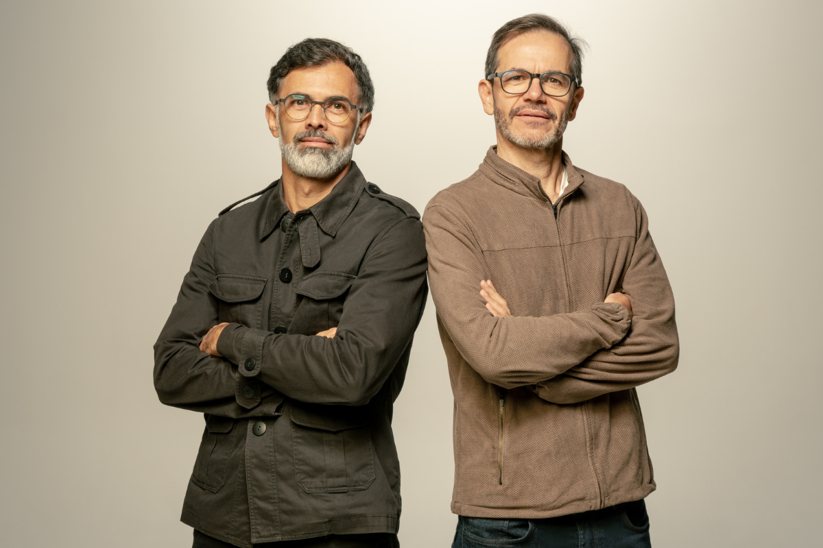 Dois homens de meia-idade, um com barba grisalha e jaqueta escura, outro com jaqueta marrom, ambos de óculos, braços cruzados, olhando para frente com expressão séria