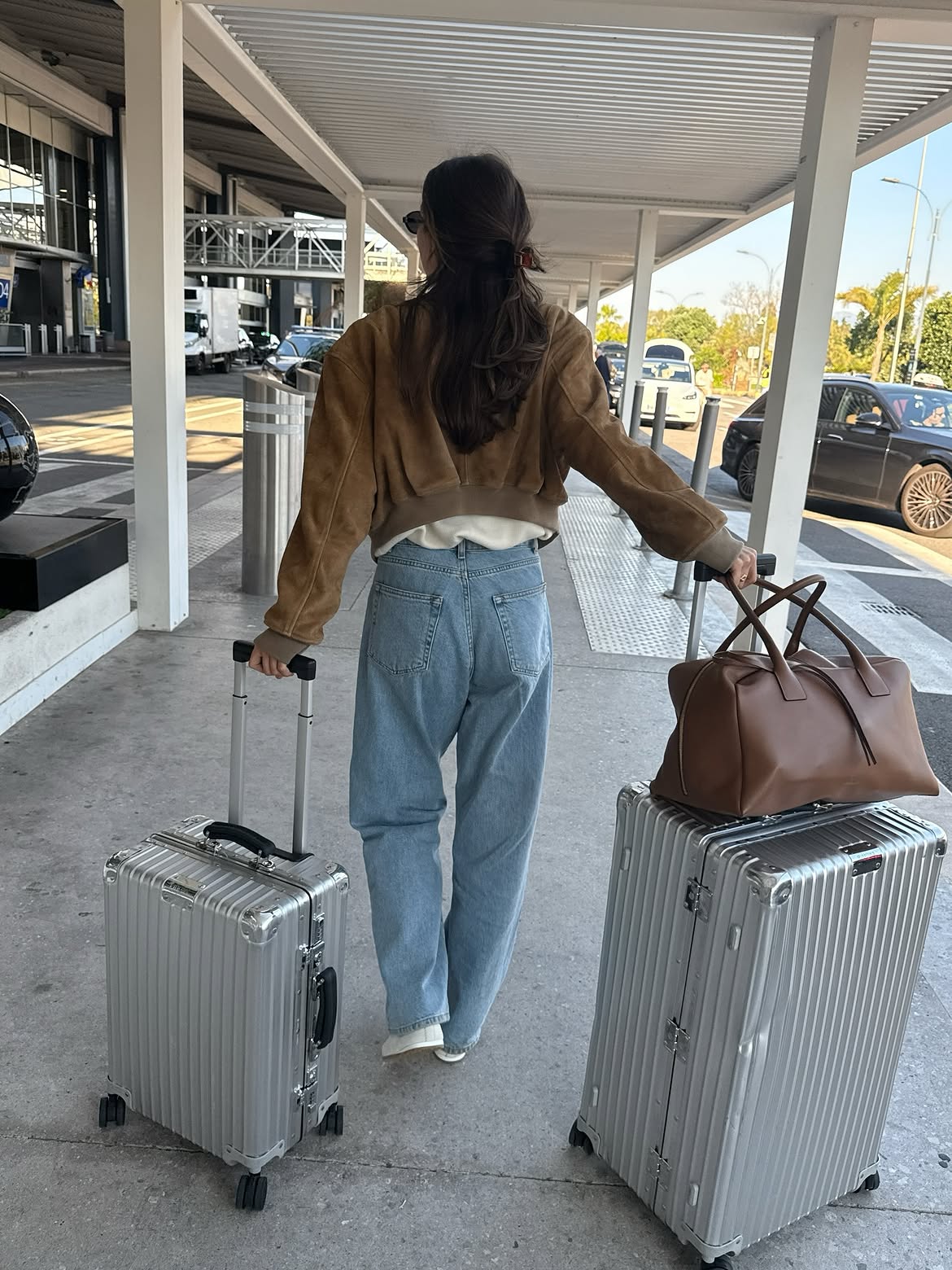 Mulher de costas, com cabelos longos e escuros, vestindo jaqueta marrom e cal&ccedil;a jeans clara, puxa duas malas prateadas e uma bolsa marrom sobre uma delas, caminhando em cal&ccedil;ada de aeroporto com carros ao fundo