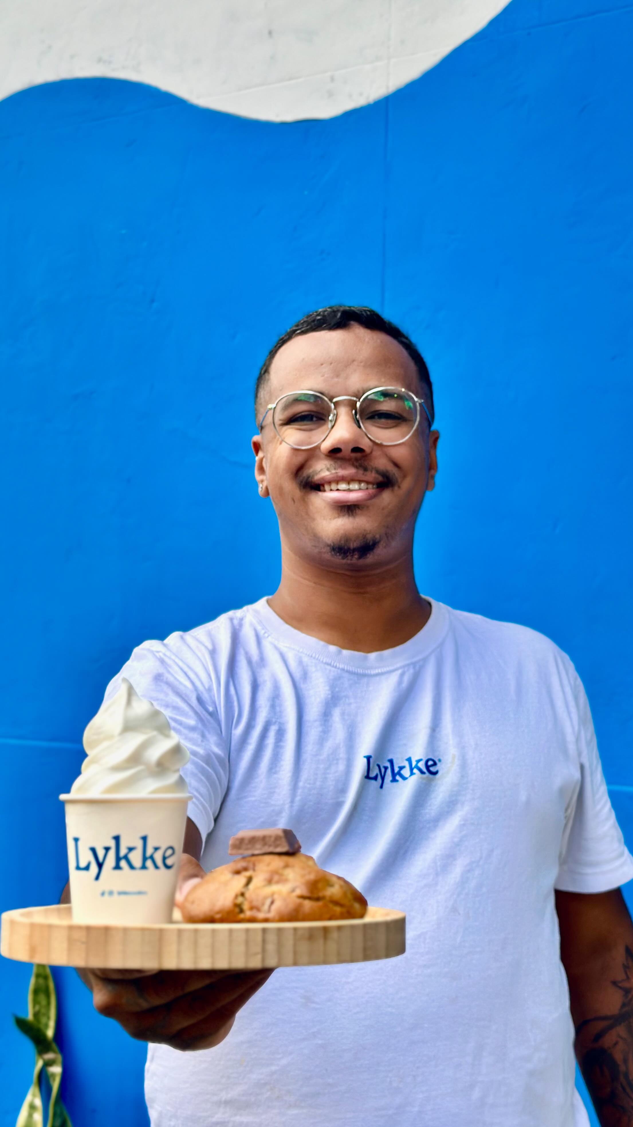 Homem negro sorridente, de &oacute;culos redondos e camiseta branca com a palavra Lykke, segurando uma bandeja de madeira com um sorvete e um cookie com chocolate, em frente a uma parede azul