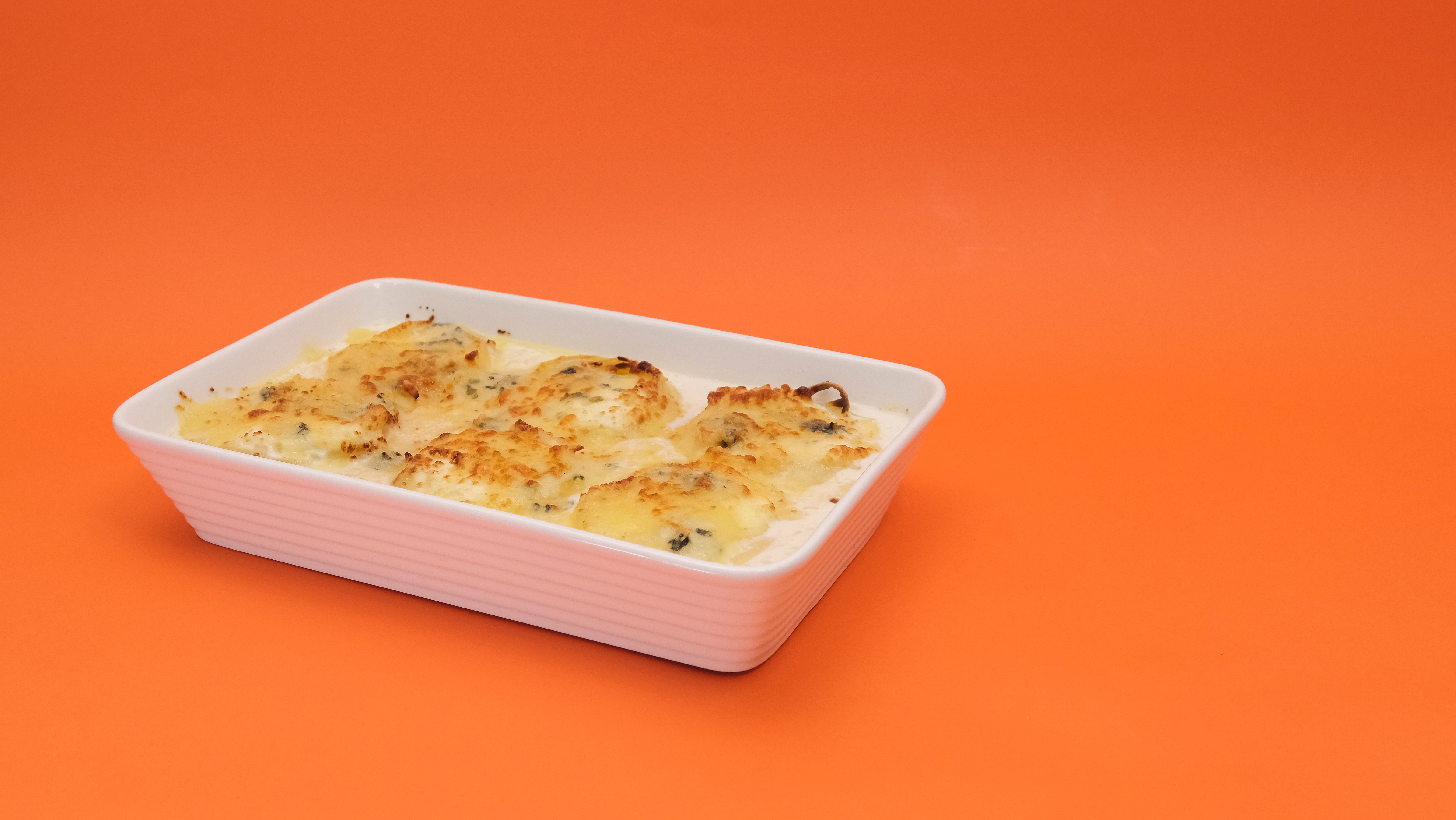 Assadeira branca retangular com nhoques gratinados em molho branco, disposta diagonalmente sobre um fundo laranja vibrante