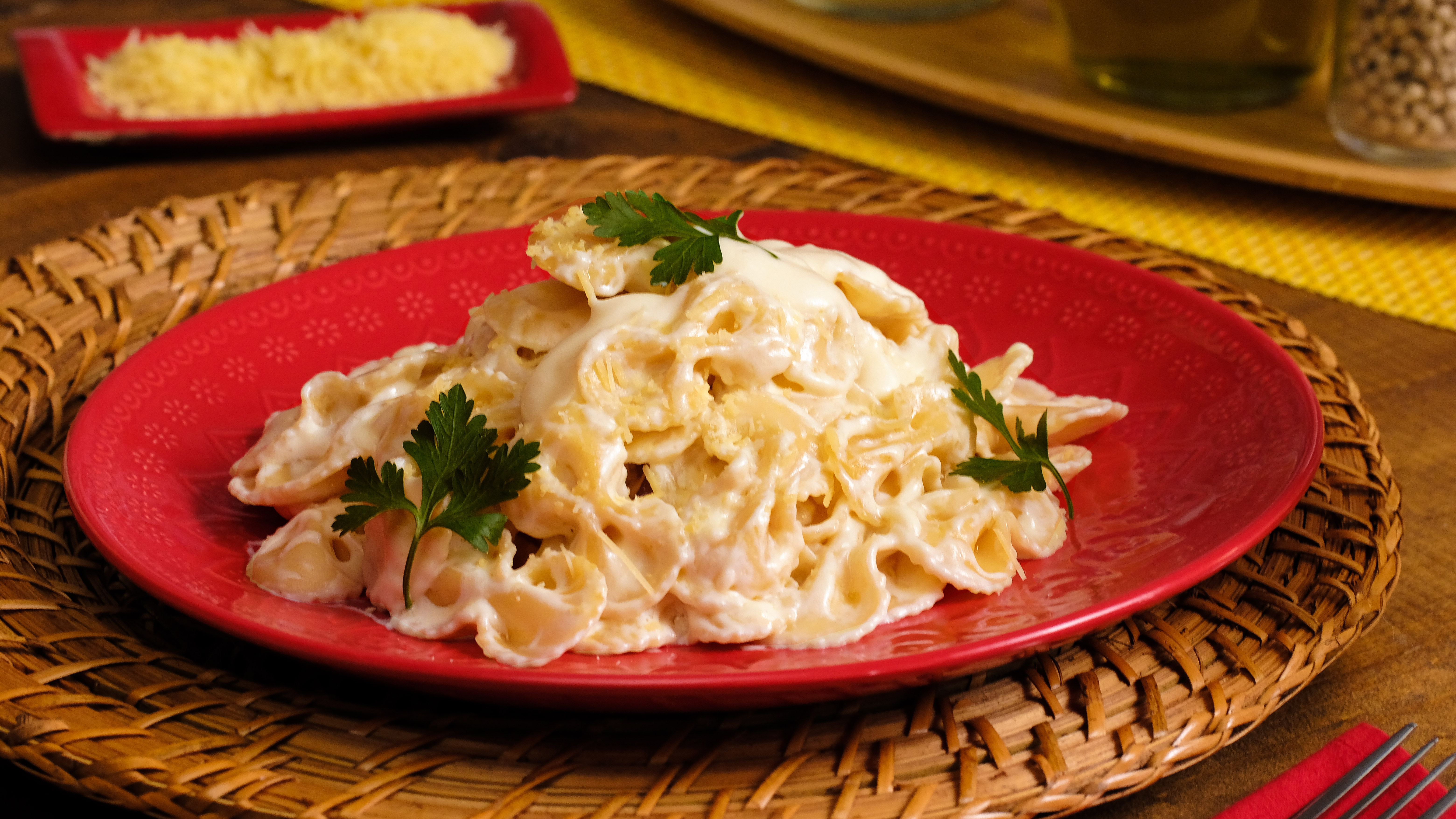 Massa tipo gravatinha com molho branco cremoso e queijo ralado, decorada com folhas de salsinha, servida em prato vermelho sobre um sousplat de palha