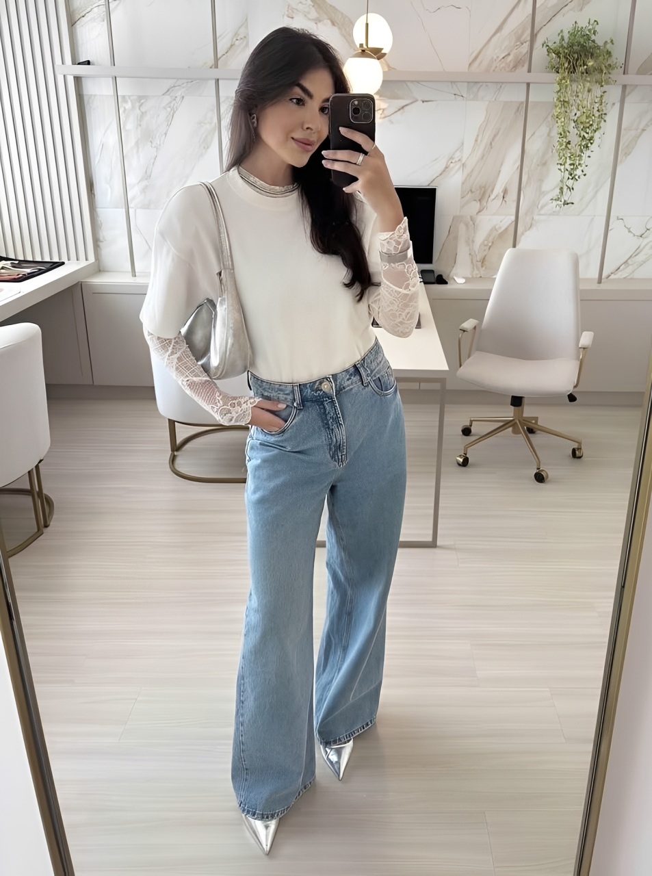 Mulher jovem de cabelo escuro, sorrindo e tirando uma selfie no espelho. Ela veste uma blusa branca de manga curta sobre uma blusa de renda branca de manga comprida, cal&ccedil;a jeans larga e sapatos prateados de bico fino. Ao fundo, um escrit&oacute;rio moderno com paredes de m&aacute;rmore, mesa branca e cadeira de escrit&oacute;rio.