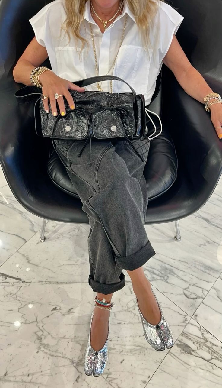 Mulher sentada em uma poltrona preta, vestindo camisa branca sem mangas, cal&ccedil;a jeans escura e sapatos prateados com bico dividido. Ela segura uma bolsa preta texturizada com dois bolsos frontais. Usa colares dourados, pulseiras e an&eacute;is, com unhas pintadas de amarelo e branco. O ch&atilde;o &eacute; de m&aacute;rmore branco com veios cinzas