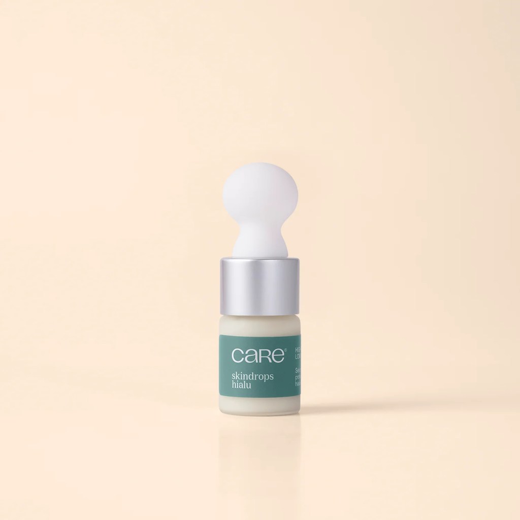 Frasco pequeno de vidro com conta-gotas, contendo um s&eacute;rum facial branco. O r&oacute;tulo verde-&aacute;gua exibe care e skindrops hialu em branco. A tampa prateada tem um bulbo branco esf&eacute;rico na ponta, sobre um fundo bege claro com reflexo do frasco