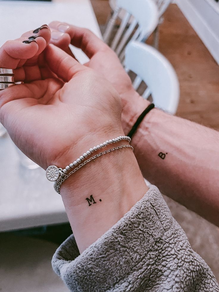 Dois pulsos lado a lado, um feminino com pulseiras prateadas e a letra M. tatuada, e outro masculino com a letra B. tatuada e uma pulseira preta