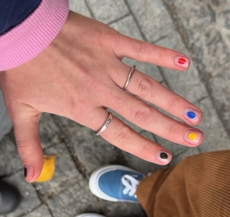 M&atilde;o esquerda de uma pessoa com unhas pintadas com bolinhas coloridas: vermelho, azul, amarelo e preto. Dois an&eacute;is prateados adornam os dedos, um liso e outro com detalhes. A pessoa veste uma blusa rosa e cal&ccedil;a marrom, com um t&ecirc;nis azul vis&iacute;vel no canto inferior direito. O fundo &eacute; um piso de paralelep&iacute;pedos.