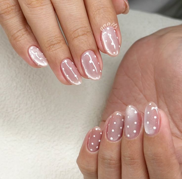 M&atilde;os femininas com unhas ovais pintadas de rosa claro e decoradas com bolinhas brancas e linhas curvas. A palavra gnails aparece no dedo anelar da m&atilde;o esquerda