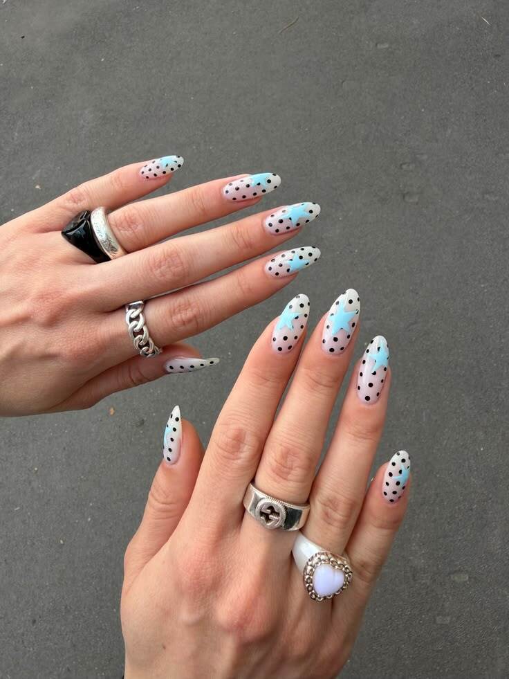 Duas m&atilde;os com unhas longas e pontiagudas, esmalte transparente com bolinhas pretas e estrelas azuis. A m&atilde;o esquerda usa an&eacute;is prateados: um preto e um de corrente. A m&atilde;o direita tem an&eacute;is prateados: um com o s&iacute;mbolo Gucci e outro de cora&ccedil;&atilde;o branco