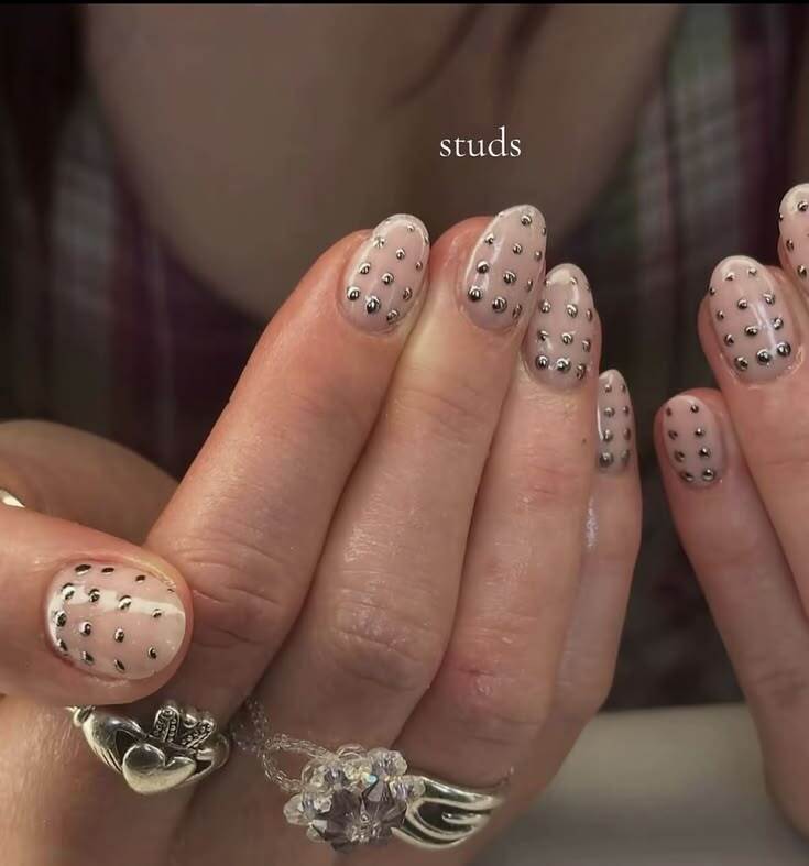 M&atilde;os femininas com unhas ovais pintadas de nude e decoradas com pequenos studs met&aacute;licos prateados. Um anel de prata com um cora&ccedil;&atilde;o e coroa e outro anel com pedras roxas adornam os dedos. A palavra studs aparece no topo da imagem