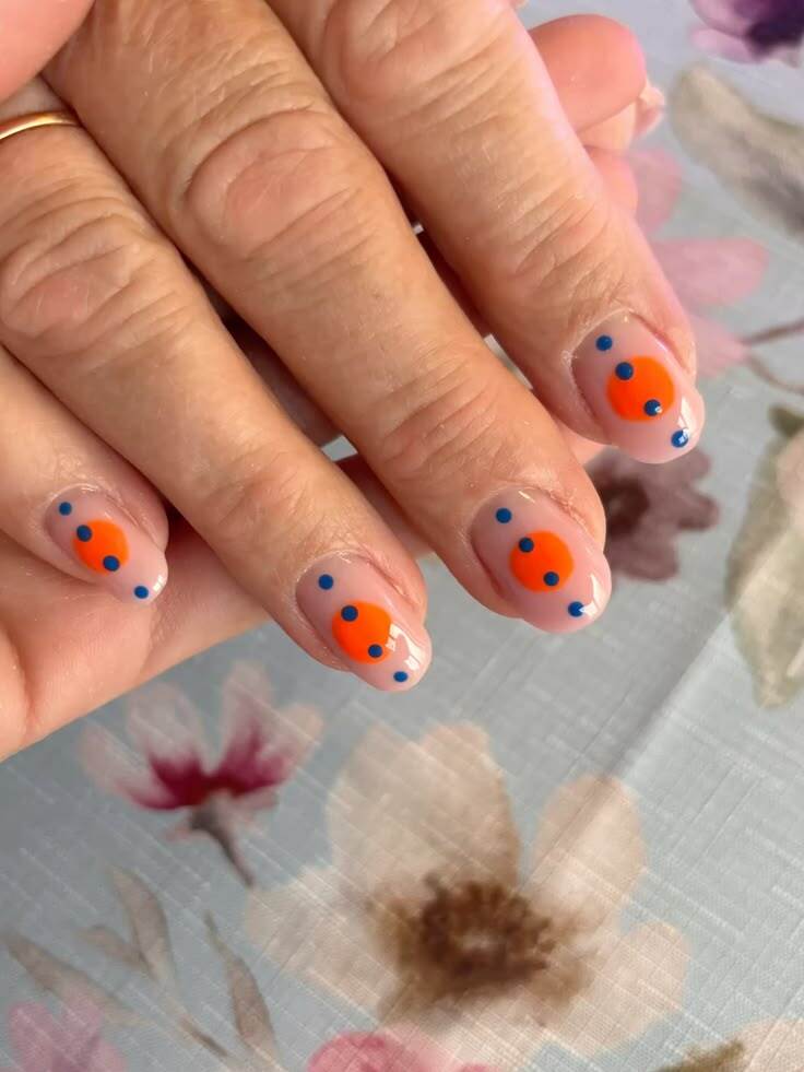 M&atilde;o feminina com unhas curtas e arredondadas, esmalte nude com um c&iacute;rculo laranja central e pequenos pontos azuis ao redor, sobre uma toalha de mesa floral azul