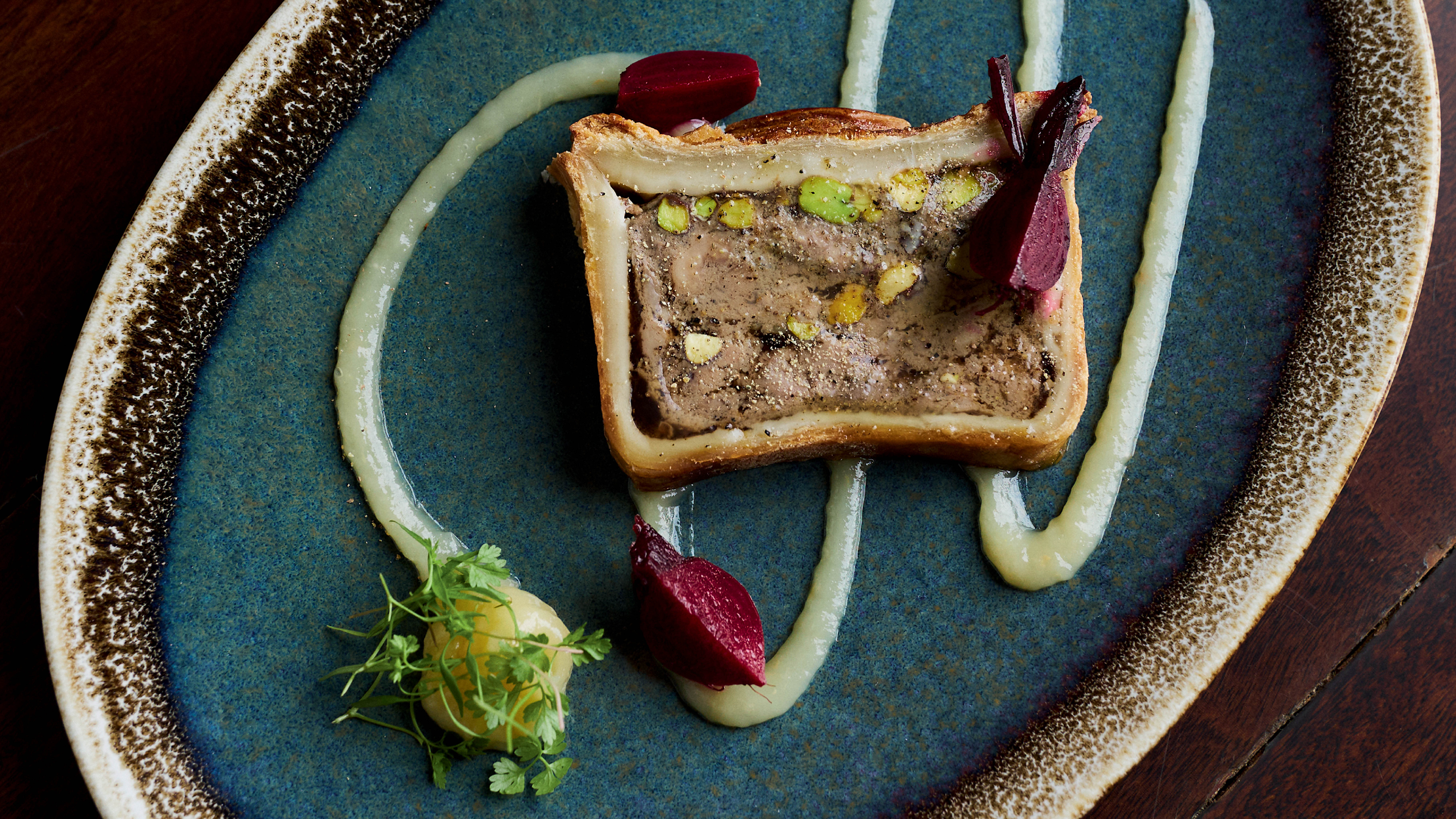 Uma fatia de patê en croute com pistaches, decorada com beterraba e um molho claro, servida em um prato azul texturizado